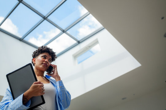 Businesswoman is holding a tablet and talking on her phone while standing under a large skylight in a modern office building, looking upwards with a serious expression - Powered by Adobe