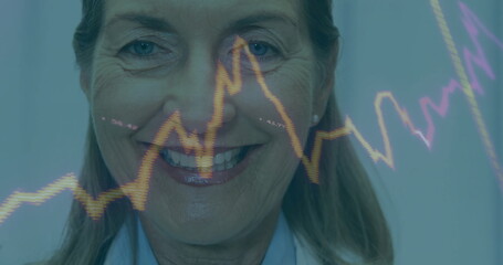 Smiling senior doctor wearing lab coat, analyzing trends in clinic with purple yellow line graphs