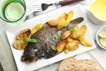 Tasty roasted flounder fish served with potatoes, basil and lemon slices on white marble table, flat lay. Homemade seafood dish