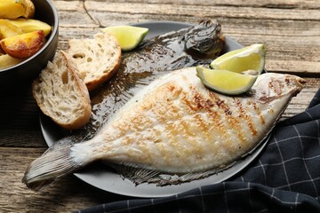 Tasty roasted flounder fish served with potatoes, bread and lime slices on wooden table, closeup....