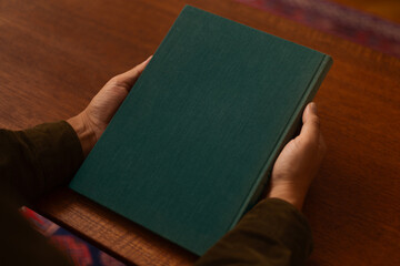 A person holds a blank linen, dark green book on a wooden table, ready to be opened. The simple,...