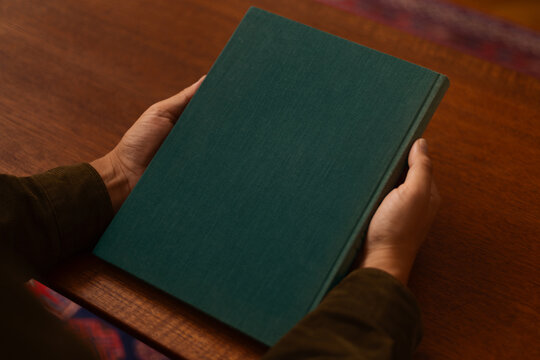 A person holds a blank linen, dark green book on a wooden table, ready to be opened. The simple, clean design and neutral setting give a sense of calm focus and classic knowledge.