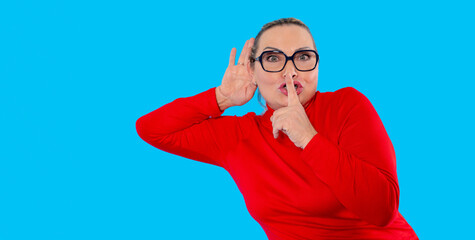 Woman in red sweater playfully gestures for silence against a bright blue background in a...