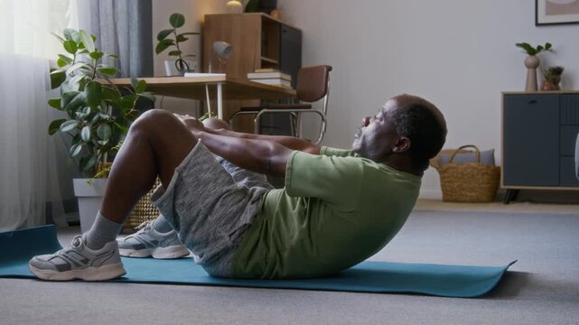 Full side shot of active elderly African American man lying supine on sports mat on floor in living room at home, performing abdominal crunch exercise with body weight while keeping fit in old age