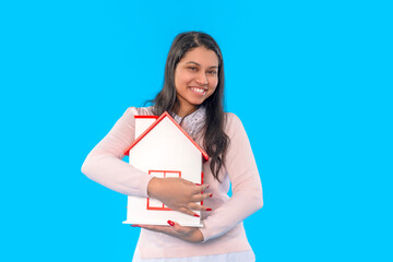 Young woman joyfully holding a miniature house against a bright blue background while expressing happiness and excitement about homeownership