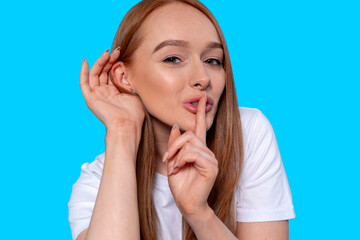 Young woman with long red hair playfully making a shushing gesture against a bright blue background...