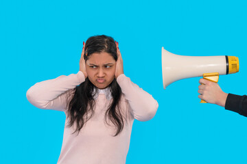 Woman covering ears while another person holds a megaphone against a bright blue background during...