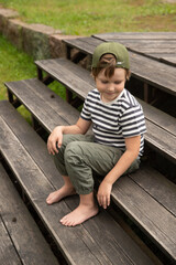 Fototapeta premium A boy sits on a wooden staircase outside in the summer. A boy laughs in a cap barefoot on a wooden platform near the lawn.