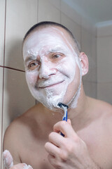 Close-up portrait of a man covered in thick shaving foam while shaving in front of a bathroom...