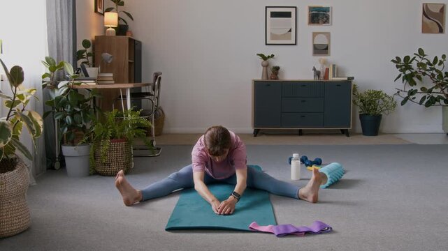 Wide shot of sporty elderly Caucasian woman in pink tshirt and blue leggings sitting on yoga mat on floor in living room, doing forward bend exercise to stretch hamstrings and lower back