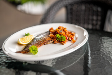 Grilled fish fillet with crispy skin served with diced vegetables and lemon wedge on a white plate....