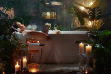 Woman enjoying a bubble bath with headphones and a tablet, surrounded by plants and candlelight.  A scene of pure relaxation and self-care.