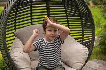 A boy sits outside in a wicker swing in summer, a boy laughs, wonders barefoot in a striped cocoon.