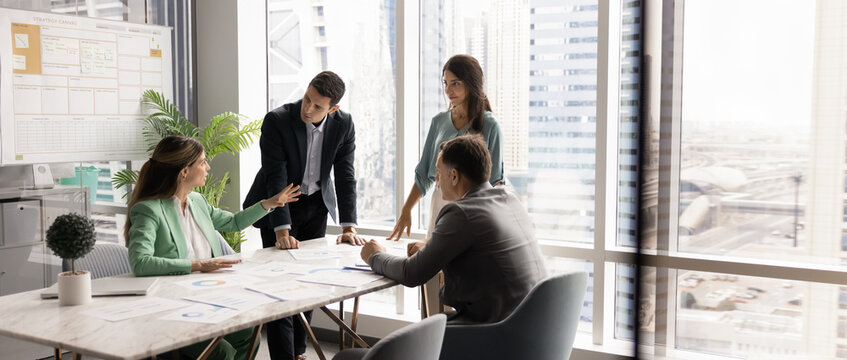 Panoramic image of four professionals negotiating, making decisions in conference room. Confident female executive leads strategic discussion at team meeting in modern office overlooking city skyline