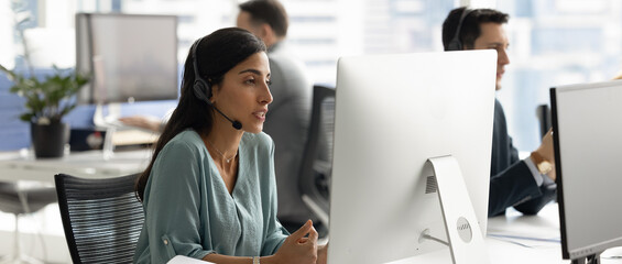 Woman wear headset speak, look at computer monitor, engaged in professional conversation, providing...