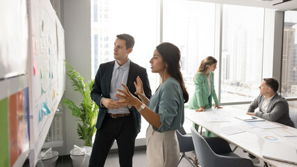 In modern office with city view, woman and man standing near whiteboard filled with sticky notes,...