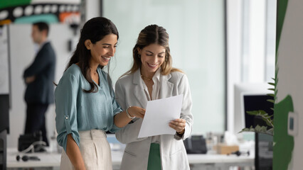 Two smiling Latina female colleagues looking at documents with positive expression, reviewing...