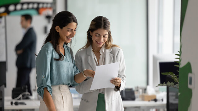 Two smiling Latina female colleagues looking at documents with positive expression, reviewing project proposal, analyzing marketing materials or designs, going over client feedback or survey results