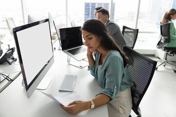 Young Latina woman deeply focused on reviewing important document, agreement or contract seated at table with modern computer with white blank mock up monitor, working in contemporary coworking office