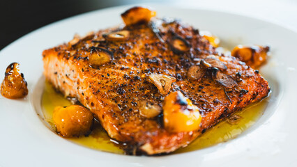 Succulent pan-seared salmon drizzled with olive oil, topped with crispy burnt garlic slices and roasted yellow tomatoes. Gourmet plating, highlighting textures and colors for food branding and culinar