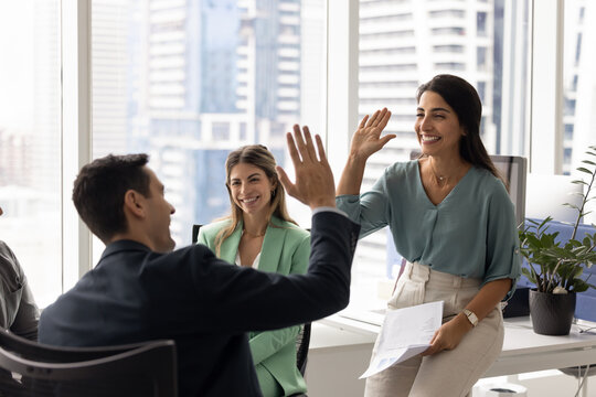 Two Latin male, female colleagues share victorious high-five during briefing meeting, celebrating shared success or breakthrough in business. Teamwork, motivation, and positive friendly work relations