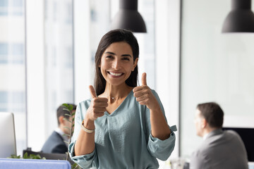 Happy Brazilian businesswoman standing in office, looking at camera, showing thumbs-up, gesture of...