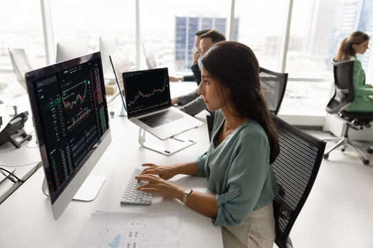 Professional Latina woman sits at desk in coworking office, focused on her work, looking at computer screen displaying financial charts and stock market data related to trading or financial analysis