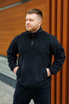 A young man in a stylish black fleece jacket stands confidently outdoors in a vibrant urban setting, smiling and embodying a trendy, carefree lifestyle that many aspire to emulate