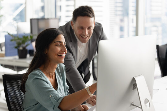 Cheerful female employee engaged in positive collaborative conversation with male colleague, smiling, points at desktop monitor, showing new program, review good sales outcome, successful work results