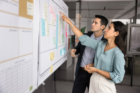 Brazilian woman and Latino man engaged in focused discussion in front of large whiteboard filled with diverse sticky notes, charts, and planning grids. Strategizing, goals reviewing and collaboration