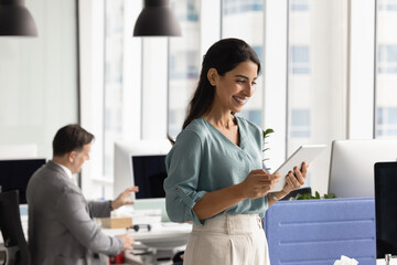 Young 30s smiling Latin woman standing in modern coworking office, holding tablet, reviewing...