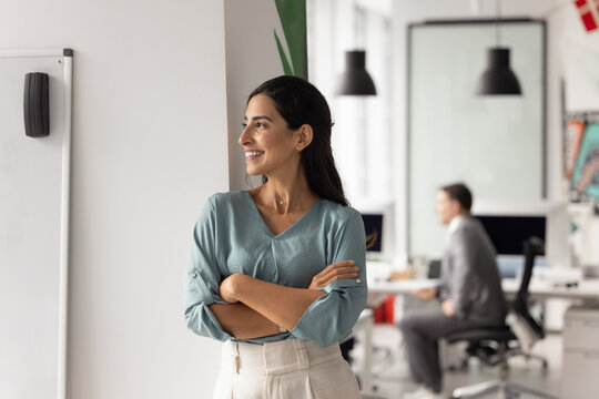 Confident Brazilian businesswoman take break stand in modern coworking office with her arms crossed, smiling, looks away thoughtfully, exuding leadership and optimism. Strategic thinking, achievements