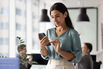 Young 30s female employee using smartphone in workplace, checking messages, reading emails or...