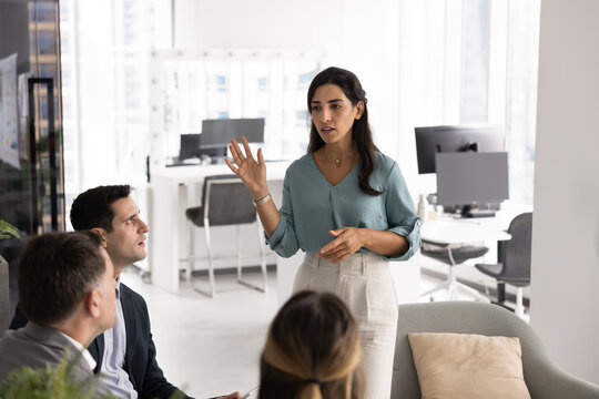 Serious Latina businesswoman speaks, stands confidently in front of small group of colleagues, lead presentation or discussion, engaged in briefing event, team meeting, collaborative strategy session