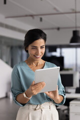 Positive Brazilian businesslady working on digital tablet device stands alone in contemporary...