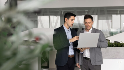 Two multiethnic men dressed in formal suits analyzing information displayed on laptop standing in...