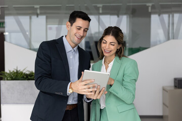 Latin colleagues standing in modern office, smiling, look at tablet device, collaborating on...