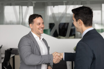 Two smiling businessmen dressed in suits standing in office, shaking hands, as gesture of mutual...