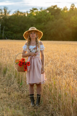 Beautiful young woman in traditional Bavarian dress and straw hat standing in golden wheat field,...
