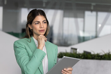 Focused Latina businesswoman holding modern digital tablet standing alone thoughtfully in modern...