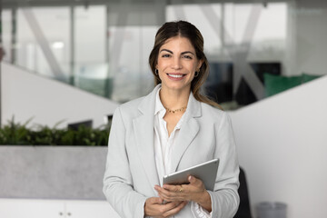 Portrait of happy Hispanic businesswoman dressed in elegant formal suit holding digital tablet,...