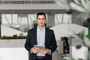 Portrait of serious Hispanic businessman dressed in formal suit holding tablet device standing in...