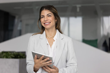 Portrait of happy Latina businesswoman stands in modern office holding digital tablet, look into...