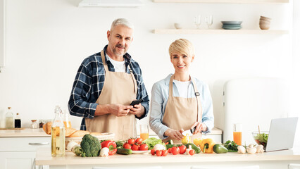 Food blogger, cooking lesson, self-isolation from covid-19 quarantine. Adult cheerful wife makes salad, man with smartphone at kitchen interior with fresh bright vegetables at table, empty space