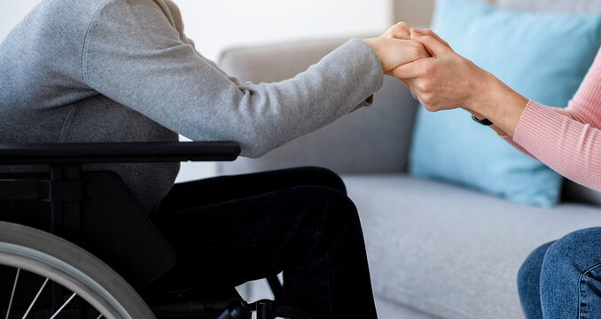 Cropped view of handicapped teenage boy in wheelchair receiving support from his mother or caregiver, holding her hands at home. Impaired adolescent feeling love from his parent indoors