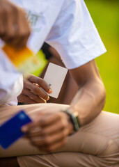 Obraz premium Black african man wearing white shirt, khaki trousers, wrist watch squating whiles holding blue card with other hands holding yellow and white cards outside .