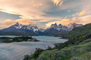 Obraz premium Majestic golden hour light illuminates the iconic Cuernos del Paine mountain range, overlooking the serene Lago Pehoé with its scenic footbridge in Torres del Paine National Park, Patagonia.