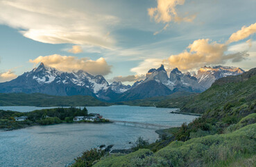 Majestic golden hour light illuminates the iconic Cuernos del Paine mountain range, overlooking the serene Lago Pehoé with its scenic footbridge in Torres del Paine National Park, Patagonia.