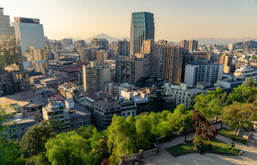 Skyline of Santiago, Chile at sunset with modern skyscrapers, historic buildings, and the Andes mountains in the distance, viewed from Cerro Santa Luc&iacute;a with lush greenery in the foreground.