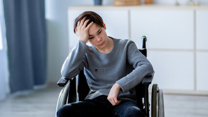 Sad disabled teenager sitting in wheelchair alone at home, copy space. Unhappy Caucasian youth with...